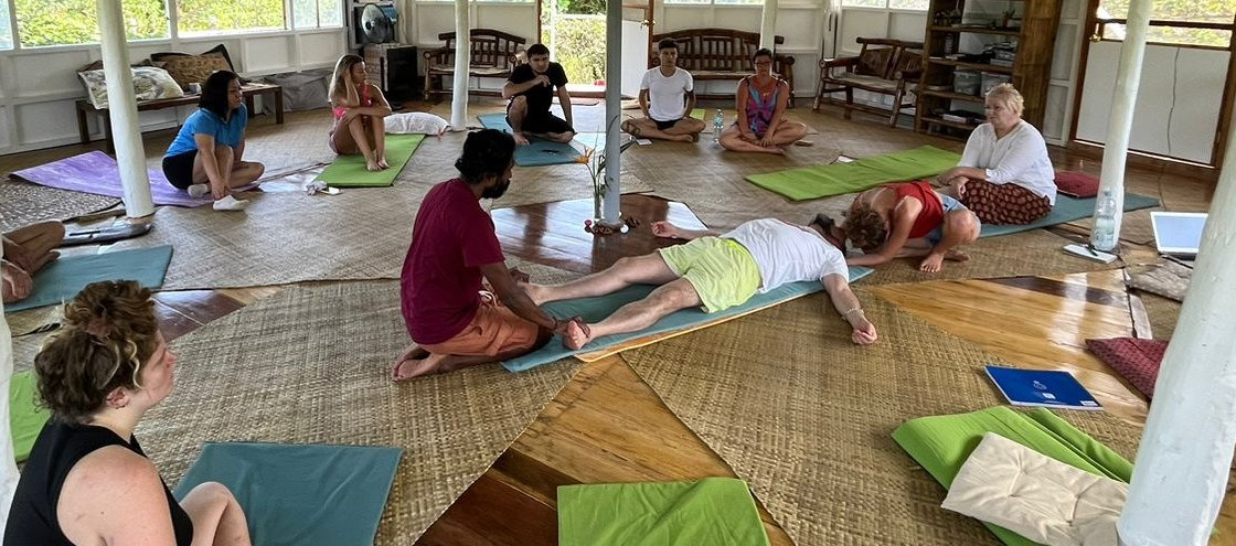 A group sitting in a circle on mats in an open wooden space, with one person lying down at the centre during a session